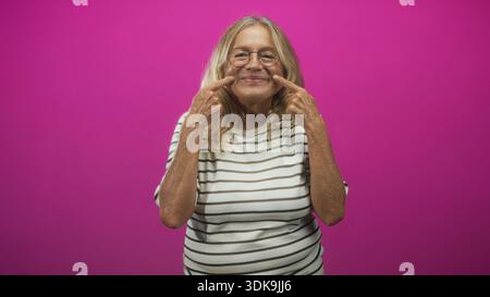 Femme senior pointant les doigts sur les joues et souriant avec les dents dans un portrait de studio sur un fond magenta ; joie ludique. Banque D'Images