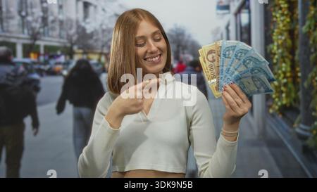 Jeune femme blonde tenant des billets de banque hongrois en forint souriant dans une rue urbaine avec un paysage urbain en arrière-plan, mettant en valeur la monnaie à l'extérieur. Banque D'Images