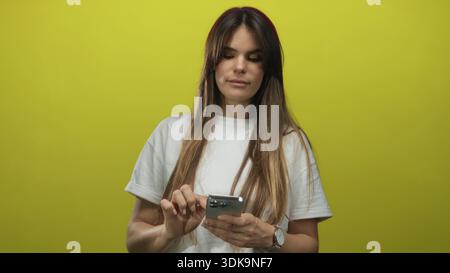 Jeune femme hispanique à l'aide d'un smartphone sur fond jaune vif portant un tshirt blanc décontracté, absorbé dans l'interaction technologique Banque D'Images