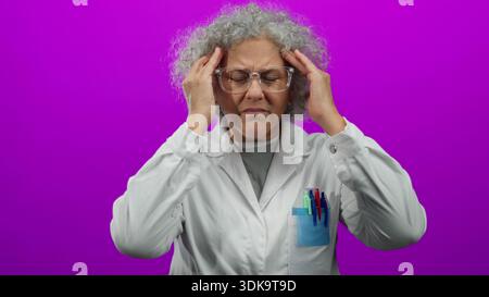 Femme en manteau blanc avec des lunettes et des cheveux gris apparaissant stressé sur un fond rose vif, isolé, avec les mains sur les tempes suggérant tête Banque D'Images