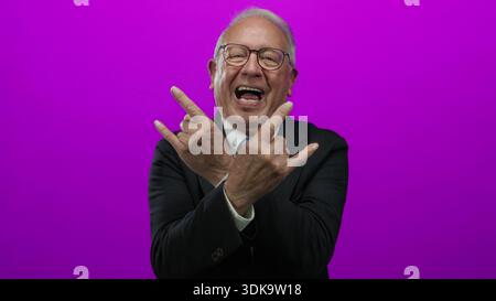 Homme senior dans un costume d'affaires faisant des gestes de main rock avec une expression ludique contre un mur de fond rose vif. Banque D'Images