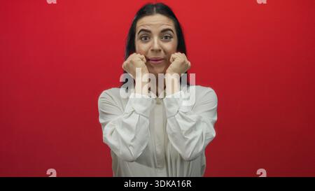 Femme exprimant l'excitation avec ses mains près du visage contre un mur rouge vif, véhiculant l'émotion intense et l'énergie, debout dans une chemise blanche, captur Banque D'Images