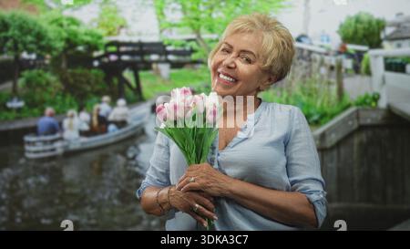 Femme hispanique senior tenant des tulipes roses, souriant tout en tenant un bouquet avec les deux mains sur la rue ; joie gratitude. Banque D'Images