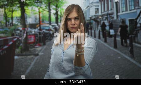 Jeune femme blonde se tient au milieu des vélos garés sur la rue pavée étroite retournant le majeur ; défi. Banque D'Images