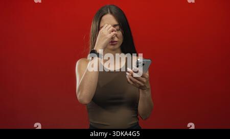 Femme à l'aide d'un smartphone, tapant l'écran avec des doigts manucurés dans un studio rouge portant un débardeur brun ; concentration. Banque D'Images