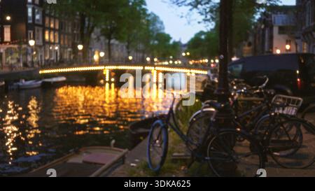 Vue floue sur le canal d'amsterdam avec des vélos au premier plan et un pont éclairé reflétant sur l'eau au crépuscule créant un effet bokeh. Banque D'Images