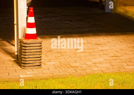 Un cône rouge et blanc se dresse sur un trottoir près de l'herbe, à côté de plusieurs poids de base lourds utilisés pour les panneaux temporaires, suggérant la configuration des travaux routiers o Banque D'Images