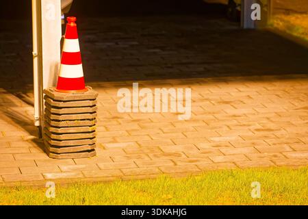 Un cône rouge et blanc se dresse sur un trottoir près de l'herbe, à côté de plusieurs poids de base lourds utilisés pour les panneaux temporaires, suggérant la configuration des travaux routiers o Banque D'Images