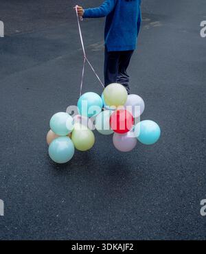 Vue arrière d'une personne marchant sur une rue asphaltée tenant un tas de ballons de couleur pastel et un ballon rouge, concept de célébration. Banque D'Images