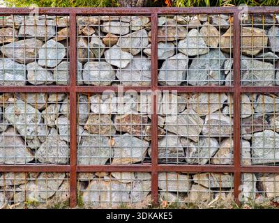 Détail architectural d'une clôture murale en gabion en pierre avec des roches grises contenues dans un cadre en treillis métallique rouillé. Banque D'Images