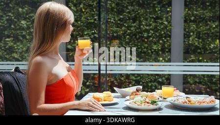 Femme buvant du jus d'orange frais pendant le petit déjeuner de luxe au restaurant en plein air. La table propose des œufs benedict, des pâtisseries et des plats sains. Hig Banque D'Images