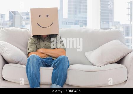 Boîte en carton avec visage souriant dessiné à la main est assis sur un canapé beige clair dans le salon. Masque, urbain, paysage urbain, lumière du jour, minimal, moderne, canapé Banque D'Images