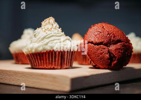 Gros plan de cupcakes en velours rouge sur une planche de bois, l'un recouvert de glaçage à la crème et de saupoudres. Banque D'Images