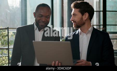 Hommes d'affaires divers collègues hommes d'affaires à la recherche d'un ordinateur portable heureux parler sans soucis discuter des résultats en ligne partenaires deux gestionnaires multiraciaux dans Banque D'Images
