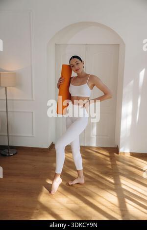 Expression faciale positive, avec tapis de yoga. Une jeune femme en tenue de fitness est dans la chambre. Banque D'Images