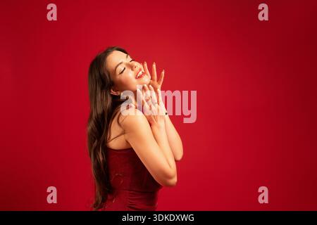 Une femme apprécie le moment, souriant les mains sur son visage dans une robe rouge devant un fond rouge. Banque D'Images
