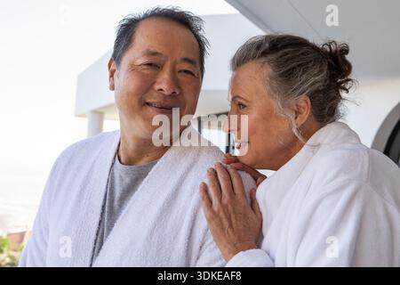 Couple senior diversifié debout sur le balcon du spa, portant des peignoirs blancs et se regardant les uns les autres. Romance, détente, bien-être, loisirs, tranquillité, architecture, horizon || modèle libéré Banque D'Images