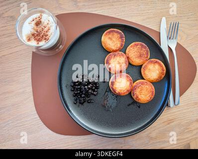 Délicieux petit déjeuner de crêpes au fromage cottage frites syrniki servies avec des myrtilles sur une assiette noire près du café avec de la mousse de cannelle. Banque D'Images
