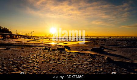 Lever de soleil doré sur une plage gelée avec des crêtes de glace et des reflets, horizon lointain de la ville et jetée à l'horizon sous des nuages spectaculaires. Banque D'Images