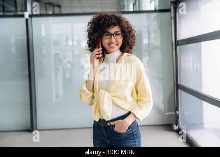Sourire en parlant au téléphone. Belle femme est debout à l'intérieur contre un mur flou. Banque D'Images