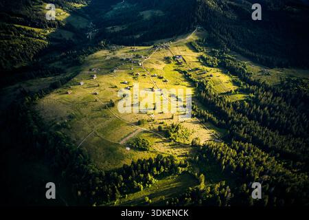 Vue aérienne de la vallée ensoleillée, parsemée de petites maisons et de routes sinueuses, entourée de forêts denses. La lumière dorée projette de longues ombres. Un paysage dynamique s'étend, se fondant dans des collines boisées. Banque D'Images