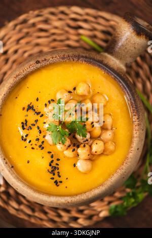 Un bol chaud de courge butternut vibrante ou de purée de citrouille servie dans un pot en céramique rustique. Le plat comporte des garnitures texturées de pois chiches entiers et fr Banque D'Images