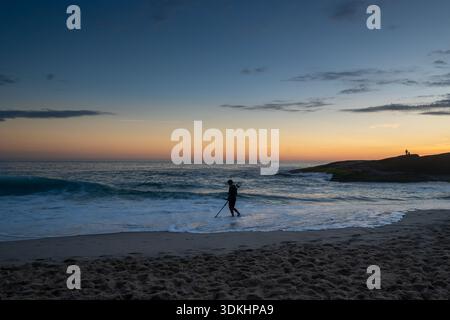 Niteroi, RJ, Brésil- 15 décembre 2025. Un homme marche avec un détecteur de métaux le long de la plage de sable d'Itacoatiara alors que le soleil se couche sur l'horizon. Banque D'Images