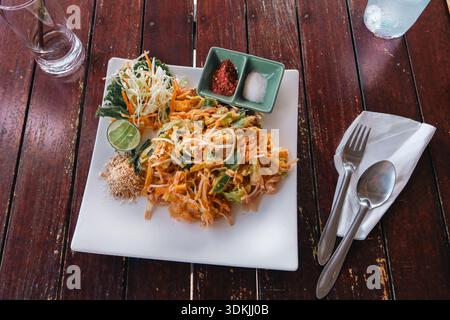 Thai Dish Pad Thai - nouilles de riz frites accompagnées de légumes, servies sur une assiette carrée blanche vue sur le dessus, sur une table en bois dans un restaurant côtier Banque D'Images
