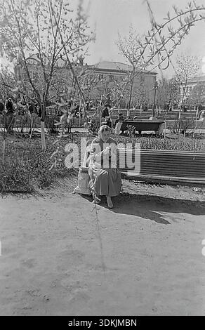 Slavyansk, Ukraine, années 1950 Une authentique photographie d'archives de Kravelis Valentina Timofeevna assise sur un banc dans un parc de la ville. La jeune femme, vêtue d'un trench à la mode et d'un foulard, regarde vers le bas. La composition est encadrée par des branches de saule floues au premier plan. En arrière-plan, il y a un bâtiment soviétique et des citoyens se relaxant sur des bancs. Ce scan haute résolution d'un négatif noir et blanc capture l'atmosphère des loisirs urbains et de la vie quotidienne paisible dans le Donbass pendant l'ère nostalgique de la vie avant la guerre. Banque D'Images