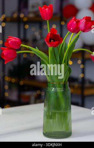 Tulipes rouges éclatantes disposées dans un vase en verre côtelé vert avec bordure dorée, lumières bokeh en arrière-plan, ambiance festive romantique Banque D'Images