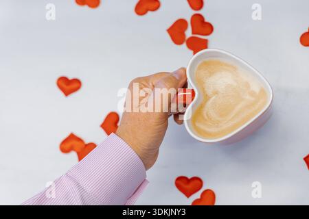 Plan horizontal grand angle au niveau des yeux des mains dans une chemise rose tenant une tasse de coeur rouge rempli de latte sur une surface blanche avec de petits coeurs rouges Banque D'Images