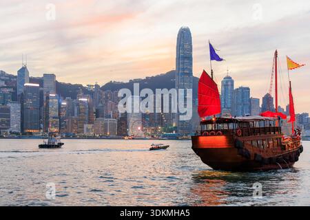 Jonque traditionnelle en bois avec des voiles rouges contre Hong Kong skyline pendant le coucher du soleil, scène urbaine emblématique Banque D'Images