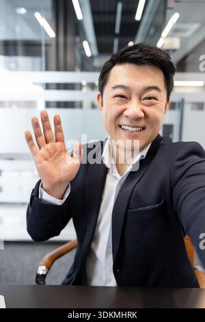 Homme d'affaires asiatique souriant et agitant lors d'une conférence vidéo en ligne conviviale depuis son bureau moderne, s'engageant avec des clients ou des collègues dans une réunion virtuelle professionnelle Banque D'Images