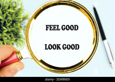 Une main de personne tient une loupe dorée avec une poignée rouge, agrandissant le texte FEEL GOOD LOOK GOOD écrit en lettres noires en gras sur un dos blanc Banque D'Images