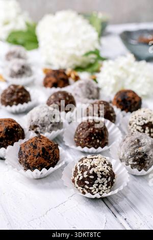 Divers boules végétaliennes crues au chocolat truffes, enrobées de poudre de cacao graines de sésame sucre en poudre placé dans des gobelets en papier sur une table en bois blanc avec hydra Banque D'Images