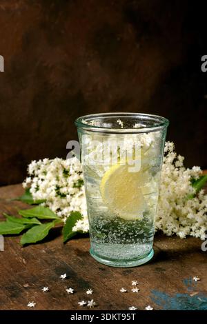 Verre eau pétillante avec tranche de citron fleurs de sureau sur table en bois rustique. Boisson rafraîchissante naturelle avec décoration de fleurs blanches Banque D'Images