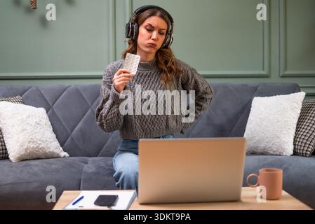Jeune femme est assise sur le lit dans le salon et ayant rendez-vous en ligne du médecin à l'aide d'un ordinateur portable. Jeune femme prenant des médicaments tout en travaillant sur lapto Banque D'Images