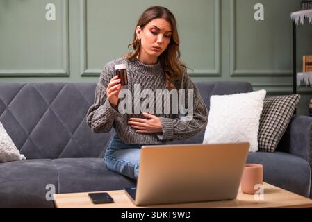 Jeune femme est assise sur le lit dans le salon et ayant rendez-vous en ligne du médecin à l'aide d'un ordinateur portable. Consultation à distance avec le médecin. Banque D'Images