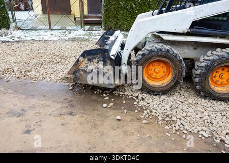 Un petit véhicule aux roues orange épand du gravier sur une surface boueuse. Un bâtiment jaune, une haie et des traces de neige sont visibles en arrière-plan. Banque D'Images