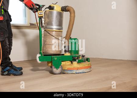 Un travailleur utilise une machine de polissage de sol vert sur un plancher en bois clair dans une pièce avec des murs blancs, une seule fenêtre et la lumière naturelle entrant. Banque D'Images