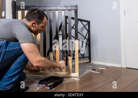 Un homme s'agenouille sur un plancher en bois recouvert de plastique, peignant un lit de bébé avec de la peinture noire. La chambre dispose d'un mur bleu clair et d'une porte blanche. Banque D'Images