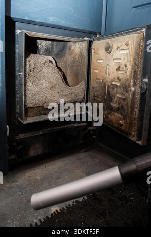 Ouvrir la porte du four métallique révèle des cendres et des résidus à l'intérieur. Une brosse d'aspirateur au premier plan suggère un nettoyage en cours sur un sol sombre. Banque D'Images