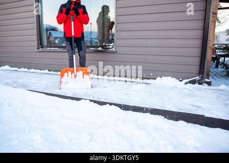 Une personne dans une veste rouge et noire tient une pelle à neige orange sur un chemin couvert de neige près d'une maison beige avec une grande fenêtre réfléchissante. Banque D'Images