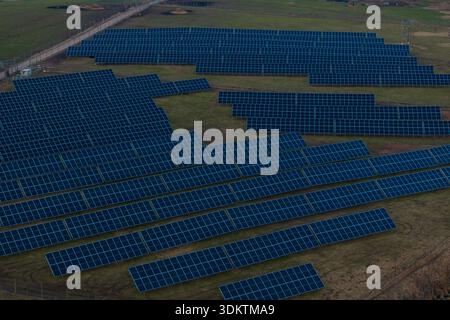 Grande ferme solaire avec des rangées de panneaux bleu foncé sur des cadres métalliques dans un champ ouvert, bordée par une clôture avec des lignes électriques visibles en arrière-plan. Banque D'Images