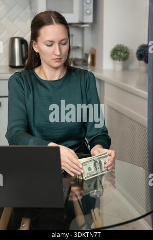 Femme d'affaires assise à un bureau en verre élégant dans un bureau à domicile, comptant une pile de billets de cent dollars tandis qu'un ordinateur portable repose à proximité, incarnant le Banque D'Images
