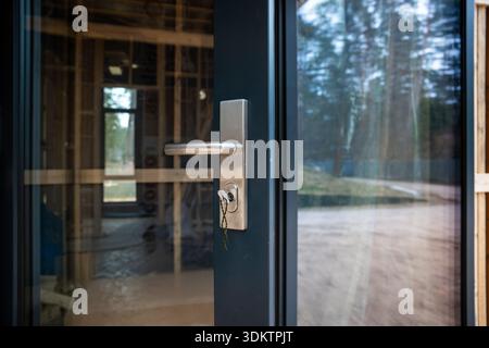 Gros plan d'une porte vitrée avec poignée métallique et clé dans la serrure, révélant l'ossature en bois et le plancher inachevé d'une maison en construction. Banque D'Images