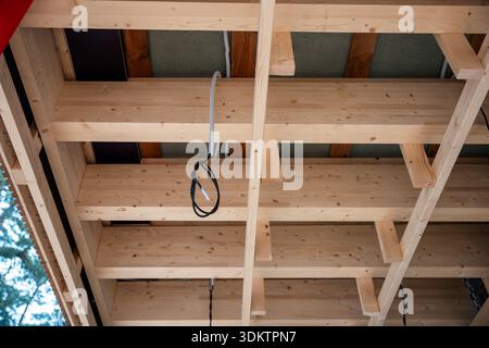 Poutres en bois apparentes et matériaux isolants dans un plafond en construction, avec câblage électrique visible. De grands arbres entourent la structure. Banque D'Images