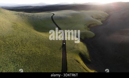 Vue aérienne d'un véhicule isolé serpentant le long d'un chemin sombre à travers des collines verdoyantes sous la douce lueur du soleil couchant, Selfoss, Islande. Banque D'Images