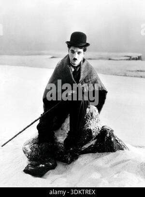 Cette photographie emblématique en noir et blanc des années 1920 montre Charlie Chaplin dans le rôle de son personnage bien-aimé, le Tramp, dans une scène de son film de 1925 « la ruée vers l'or ». Chaplin est représenté emballé dans une couverture ou un châle, assis solennellement sur un rocher dans un paysage enneigé, incarnant l'humour mélancolique de sa célèbre personnalité. Banque D'Images