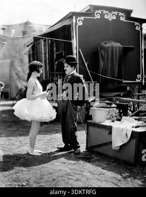 Charlie Chaplin dans le rôle de son personnage emblématique « The Tramp » sourit à une ballerine, jouée par Merna Kennedy, du film de 1928 « The Circus ». Ce noir et blanc capture encore un moment intime entre les deux personnages dans un décor de coulisses ou de foire, avec un wagon décoré en arrière-plan. Banque D'Images